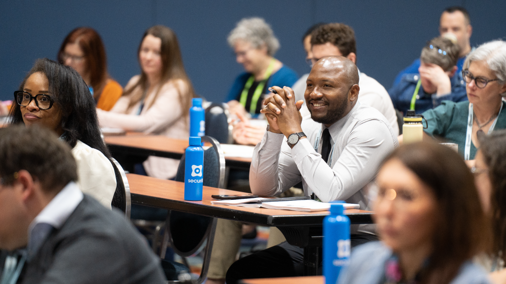 People sitting at desks attending a training seminar. In the center foreground, a young Black man wearing a tie is sitting with his elbows on the desk, smiling.