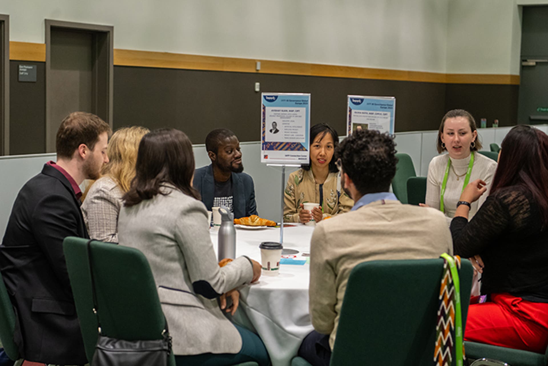 Group of attendees seated around a round table during a networking or discussion session. Table has coffee cups and informational signs.