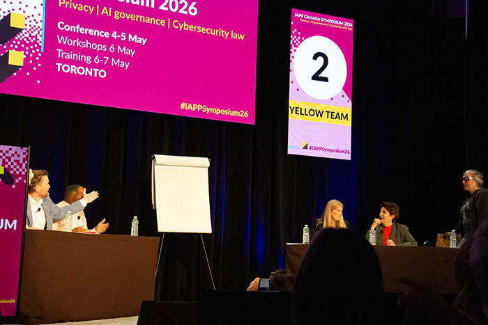 Panel discussion on stage at IAPP Canada Symposium 2026 with speakers seated at tables, a flip chart at center, and large screens displaying event details and “Yellow Team” signage.