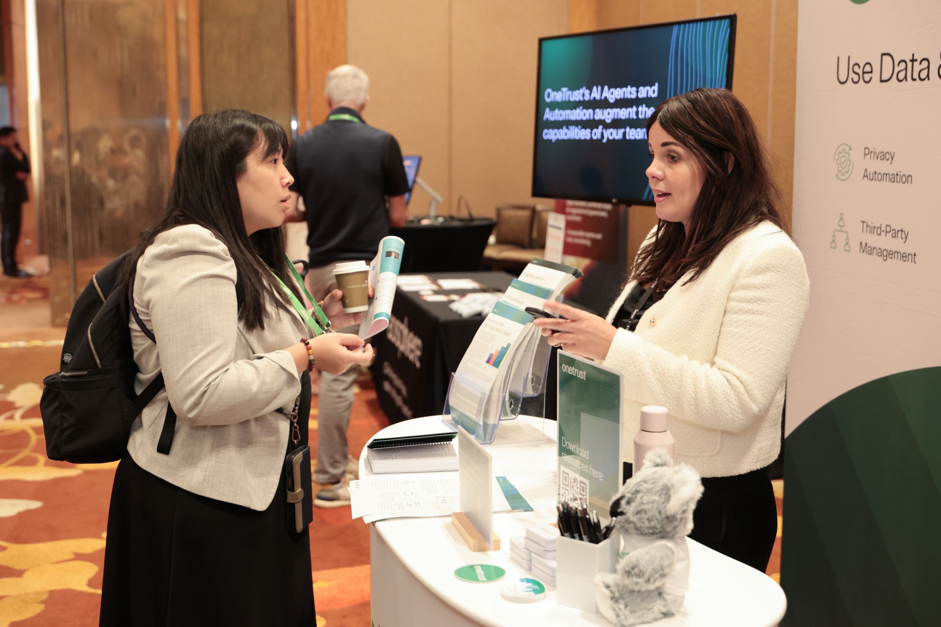 Two attendees talking at an exhibit booth displaying privacy and data management materials. The booth features brochures, pens, and a plush koala on the counter, with a banner reading “Use Data” and topics like Privacy Automation and Third-Party Management. A screen in the background shows text about AI agents and automation.