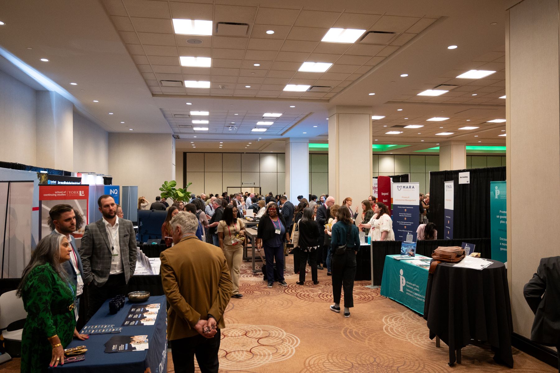 Busy conference exhibit hall with multiple booths displaying banners and promotional materials. Attendees are standing and walking throughout the space, engaging in conversations near tables covered with brochures and business cards. The room has beige walls, patterned carpet, and bright overhead lighting.