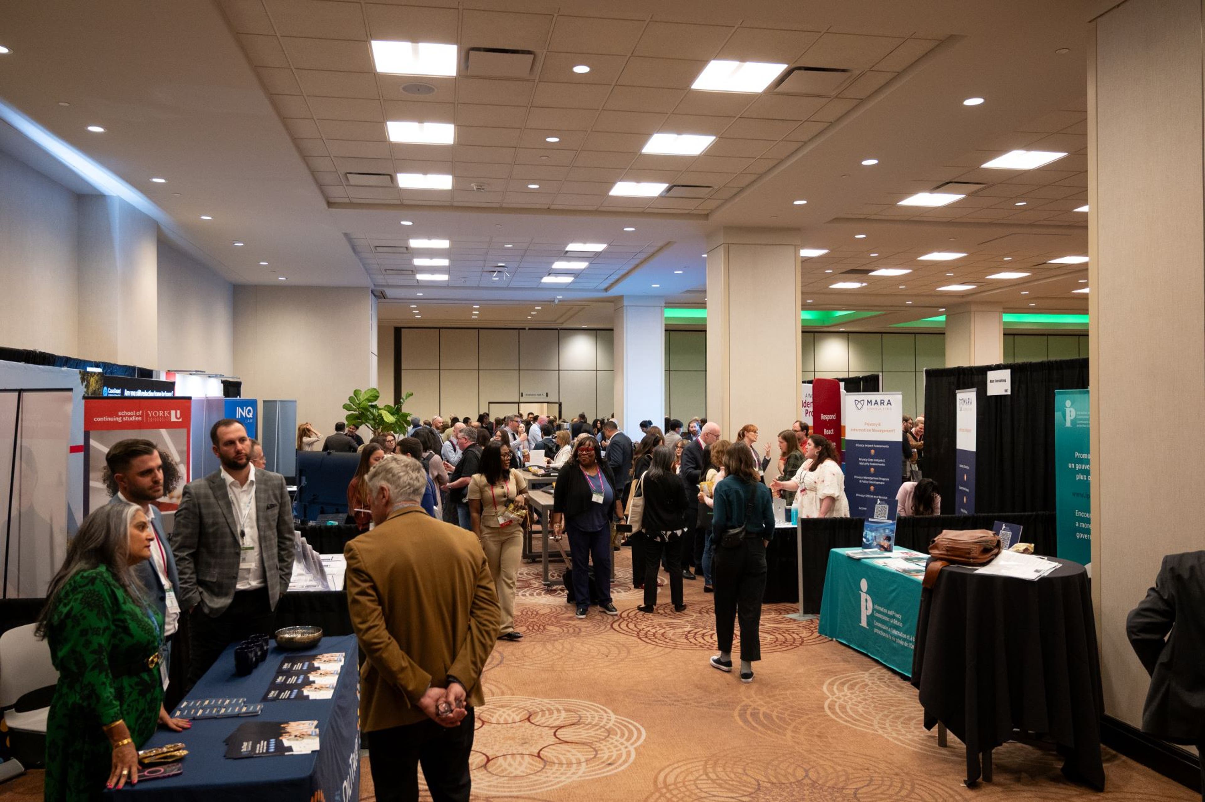 Busy conference exhibit hall with multiple booths displaying banners and promotional materials. Attendees are standing and walking throughout the space, engaging in conversations near tables covered with brochures and business cards. The room has beige walls, patterned carpet, and bright overhead lighting.