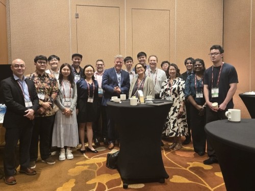 A group of conference attendees stands together around a high-top table in a networking area at an event venue. Coffee cups and event badges are visible, and the group is gathered for a posed photo in front of a neutral conference room backdrop.