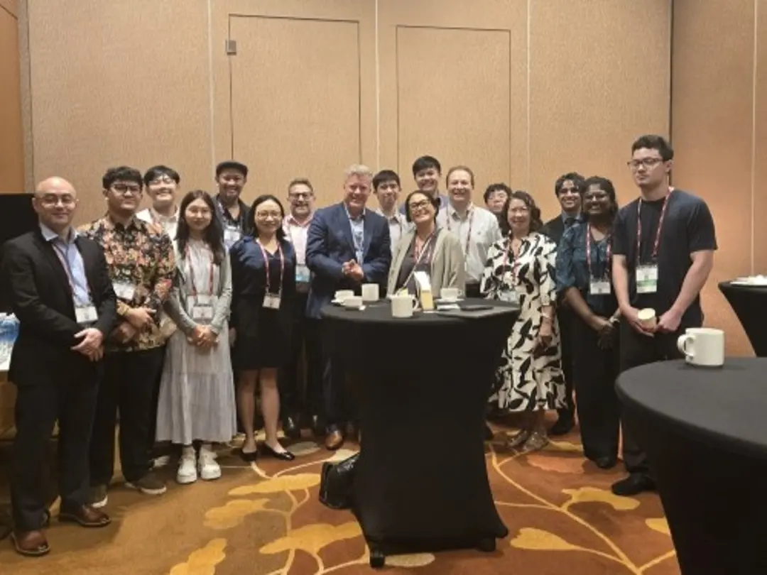 A group of conference attendees stands together around a high-top table in a networking area at an event venue. Coffee cups and event badges are visible, and the group is gathered for a posed photo in front of a neutral conference room backdrop.