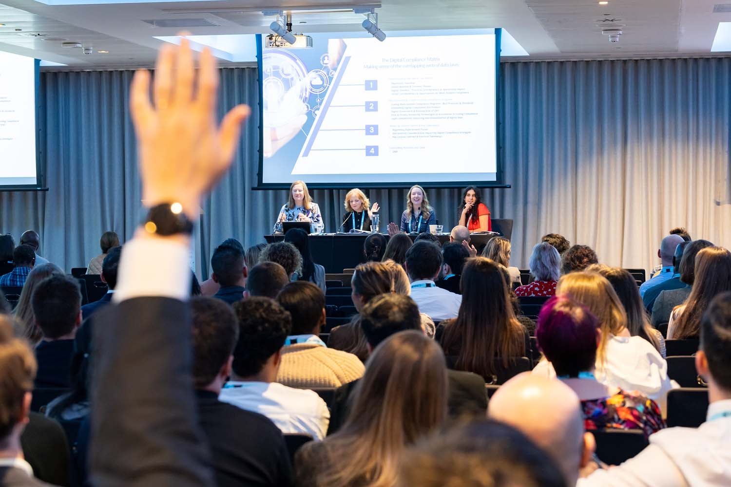An audience listens to a panel discussion at the front of a conference room while one attendee raises a hand to ask a question. A presentation slide is projected behind the panelists.