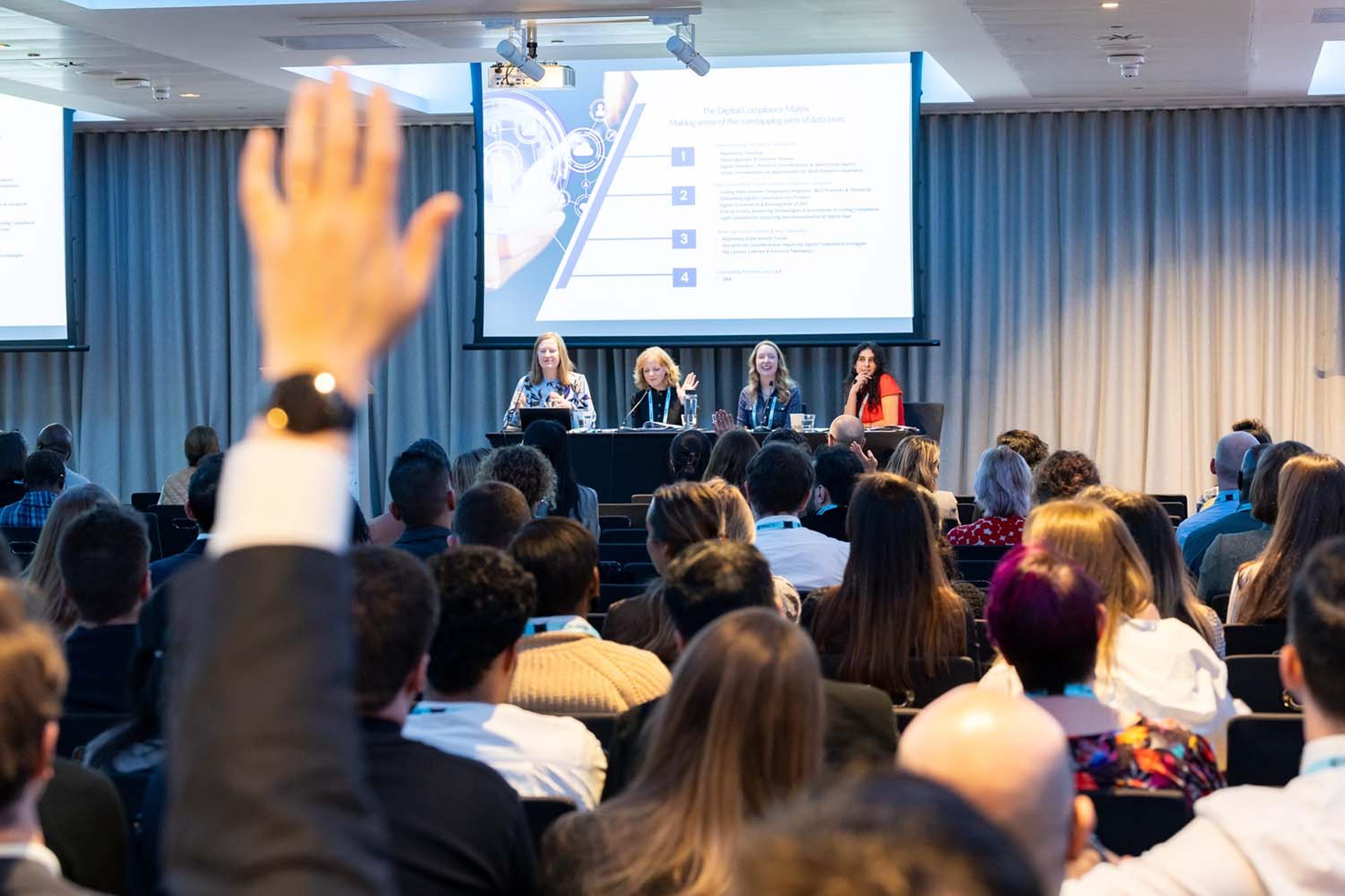 An audience listens to a panel discussion at the front of a conference room while one attendee raises a hand to ask a question. A presentation slide is projected behind the panelists.