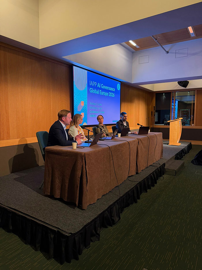 Another panel discussion takes place on a raised stage where four speakers sit at a table with microphones and open laptops. A large presentation screen behind them displays IAPP AI Governance Global Europe 2026 along with event dates and location, while a podium stands off to the side.