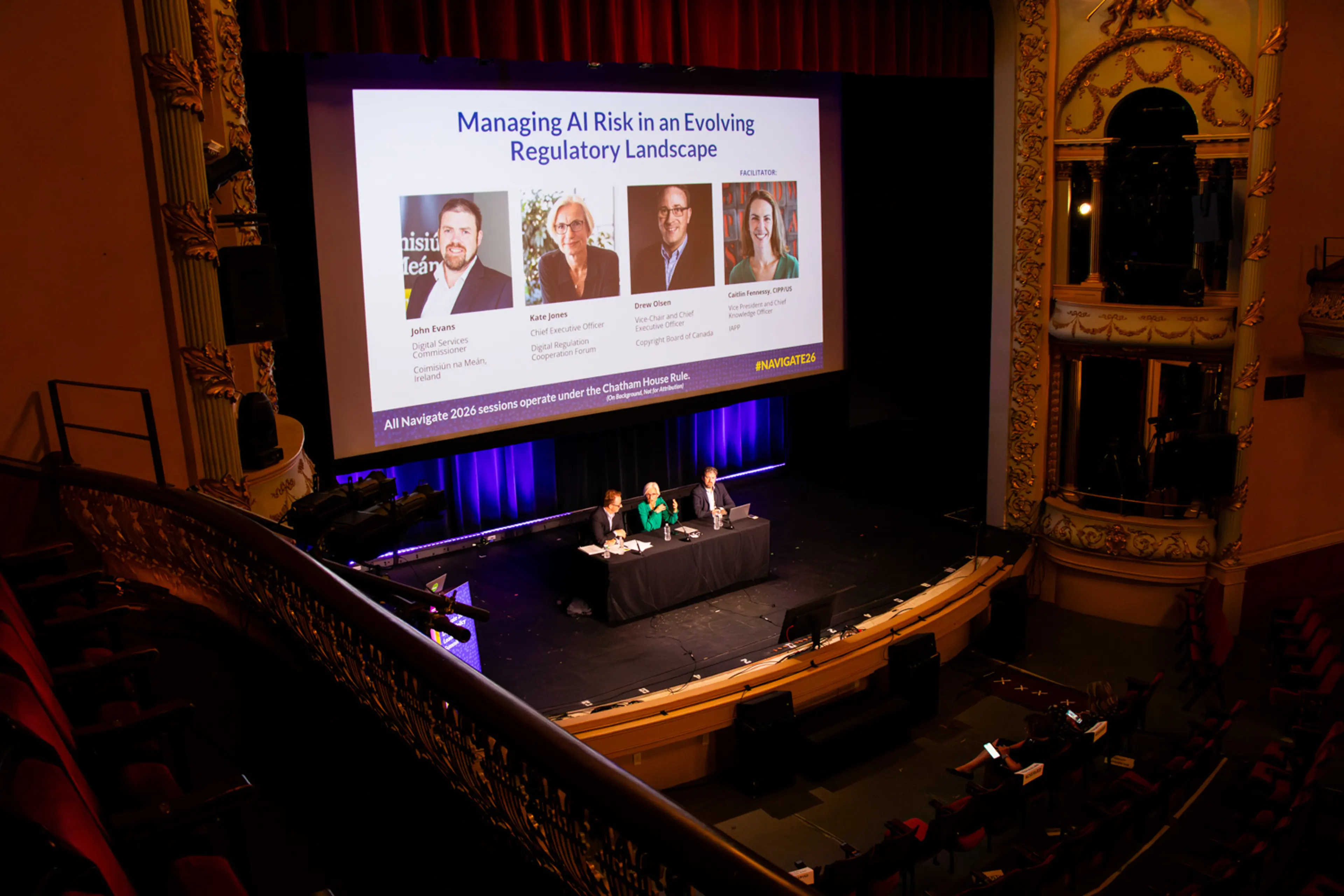 Viewed from an upper balcony, an ornate theater auditorium hosts a conference session as panelists sit on stage beneath a large screen titled “Managing AI Risk in an Evolving Regulatory Landscape,” surrounded by decorative architectural details and softly lit seating.