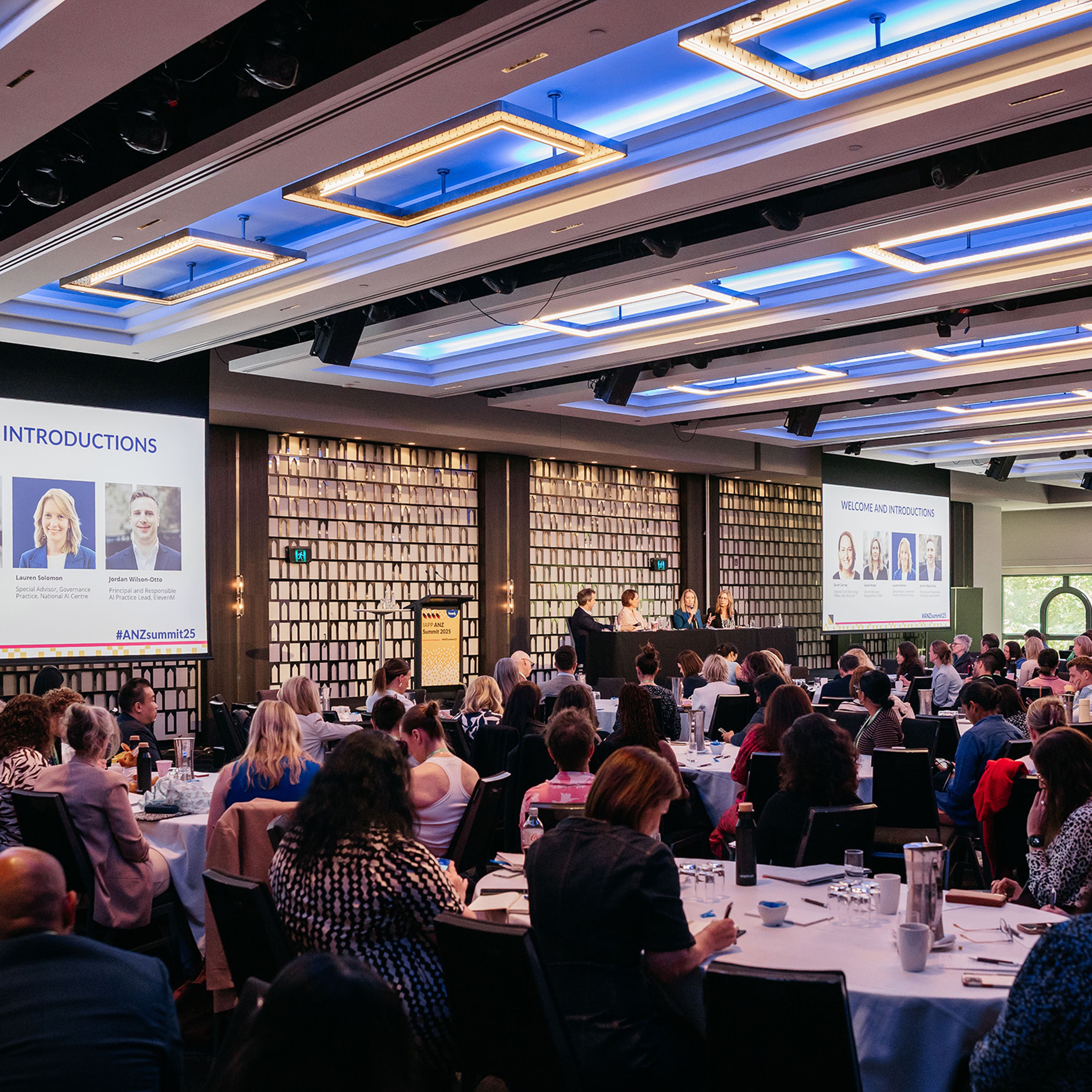 Large conference room with attendees seated at round tables, two screens displaying “Welcome and Introductions.”