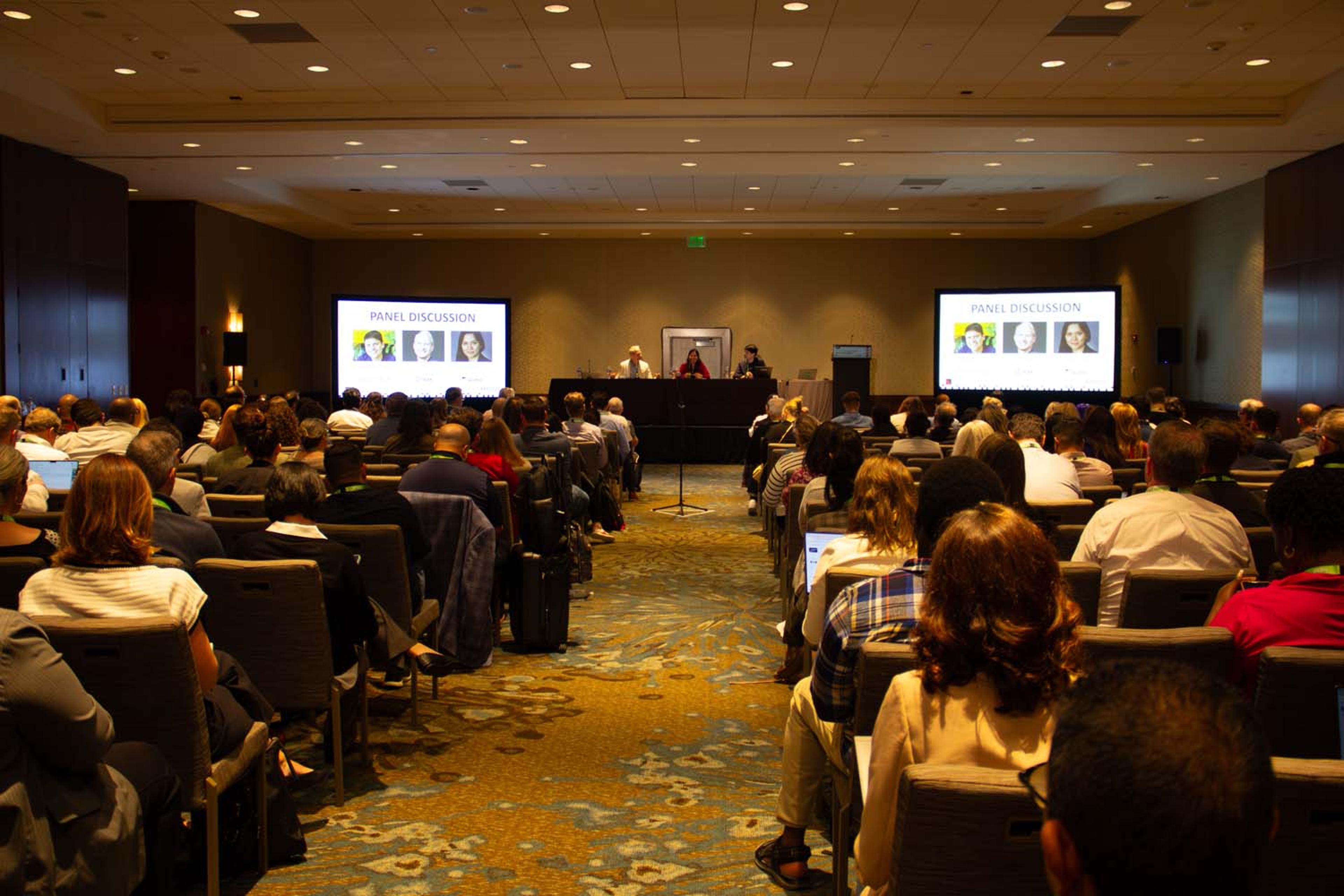 A panel of speakers sits behind a long table with microphones and water bottles, engaged in discussion as one panelist speaks and the others listen attentively.