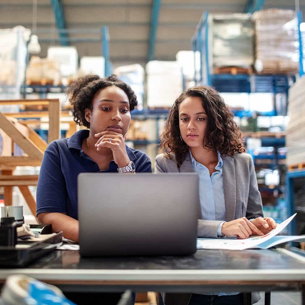 two women surrounded by boxes in warehouse looking at a laptop reviewing their inventory