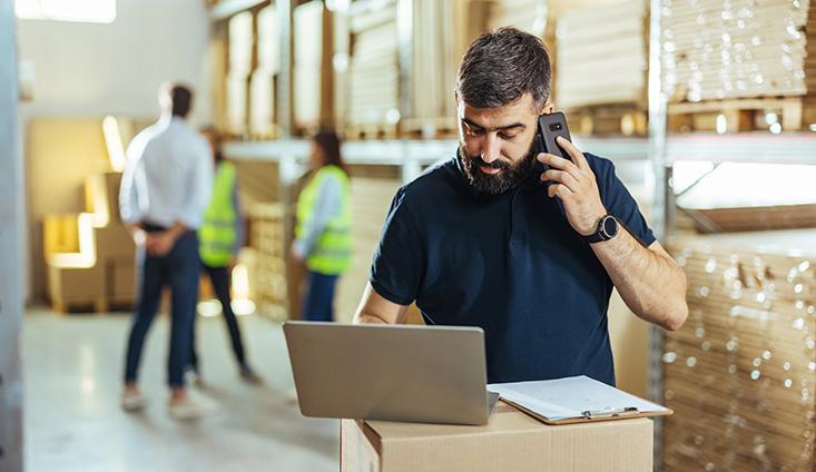 man on a phone in a warehouse with a laptop and clipboard in front of him