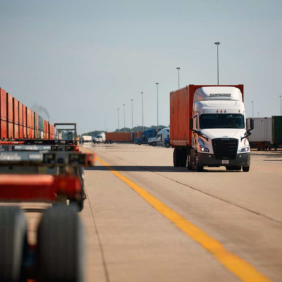 Schneider truck hauling an orange intermodal container next to a train and chassis