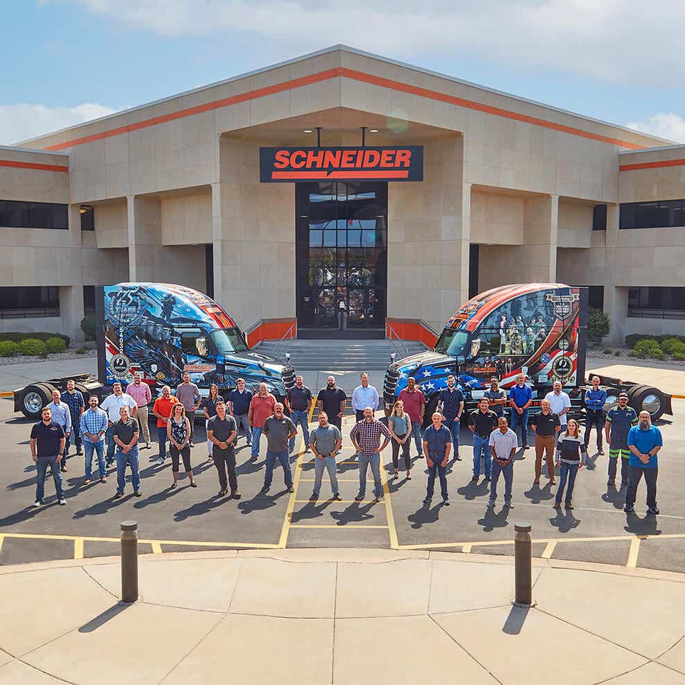 Over 30 Schneider office and shop associates with military experience standing in front of two Ride of Pride trucks, parked outside Schneider's corporate office