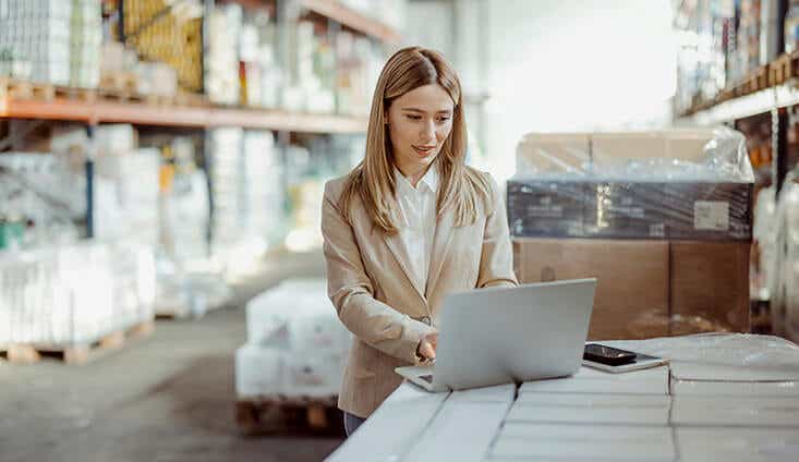 woman using a laptop in a warehouse with boxes and pallets in the background