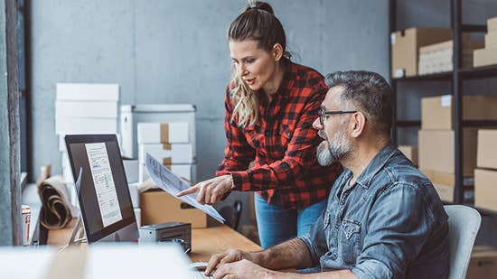 third_party_logistics.jpg A woman and man looking at a logistics document in a room surrounded by boxes.