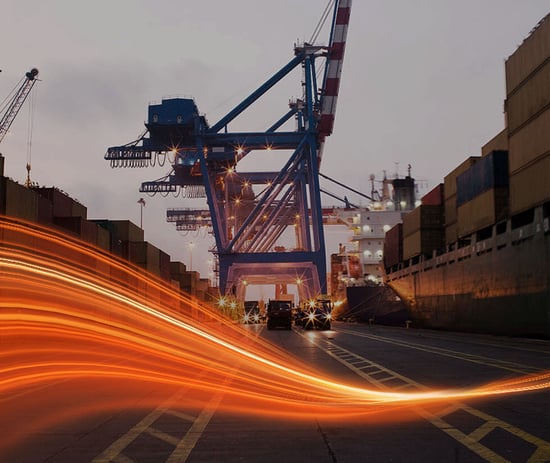vehicles driving down the center of a shipment port with cargo containers on both sides