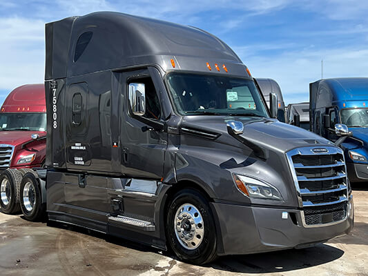A dark gray semi-truck is parked outdoors, featuring a streamlined design and prominent grille, with other vehicles in the background. The truck has orange clearance lights on top of the cab, and the numbers “758088” are printed on the side of the cabin.