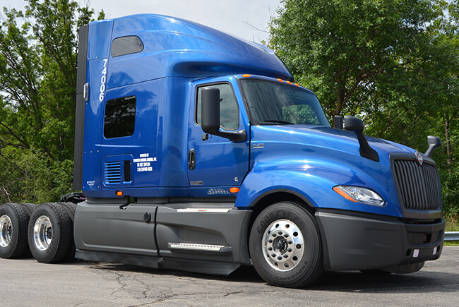 A sleek blue semi-truck parked outdoors against a green backdrop. Its aerodynamic design suggests long-haul journeys. The elevated cabin and robust wheels indicate durability. 