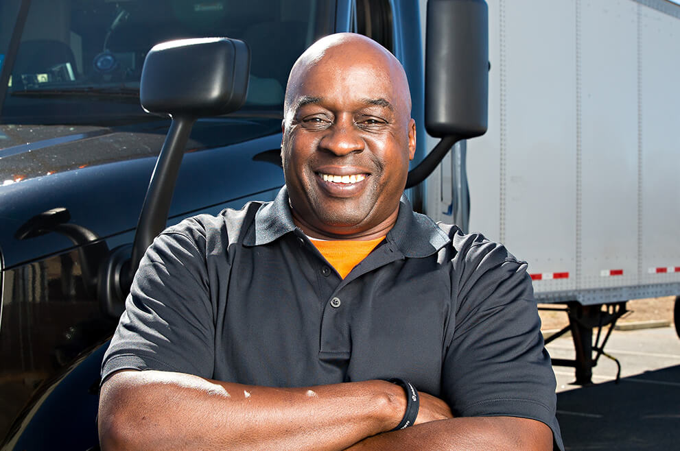 An owner-operator smiles with arms crossed confidently as he poses in front of his truck