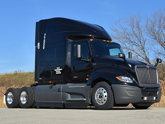 A sleek, modern, dark blue semi-truck parked outdoors under a clear sky. Streamlined design with large side mirrors and prominent front grille.