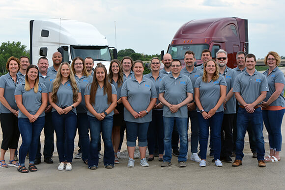 A group of people in grey shirts standing between two semi-trucks.