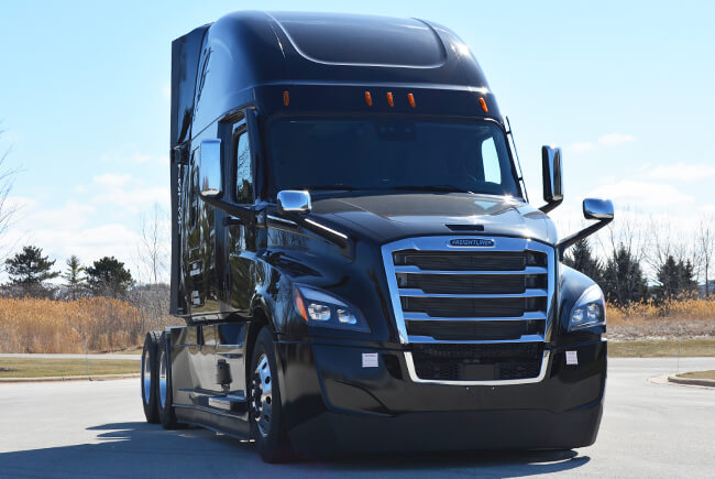 A sleek, modern black Freightliner truck parked outdoors under a partly cloudy sky. Its aerodynamic design and large side mirrors suggest efficiency and visibility.