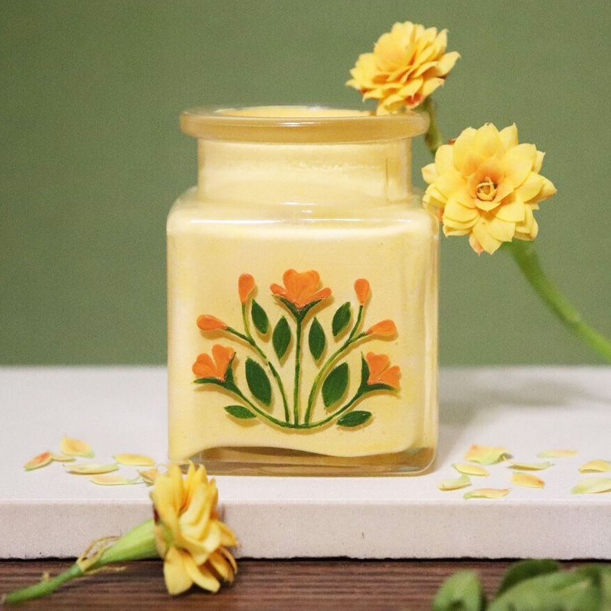 A scented candle in a yellow jar with orange flowers painted on the outside.