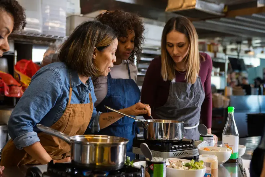 the woman demonstrates how to make a recipe to three people in her restaurant’s kitchen