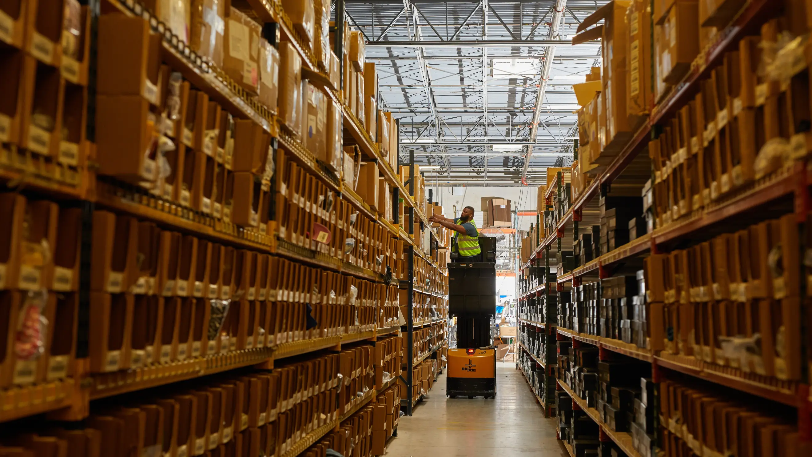 A warehouse employee uses a lift to reach items on the top shelf of a pallet rack