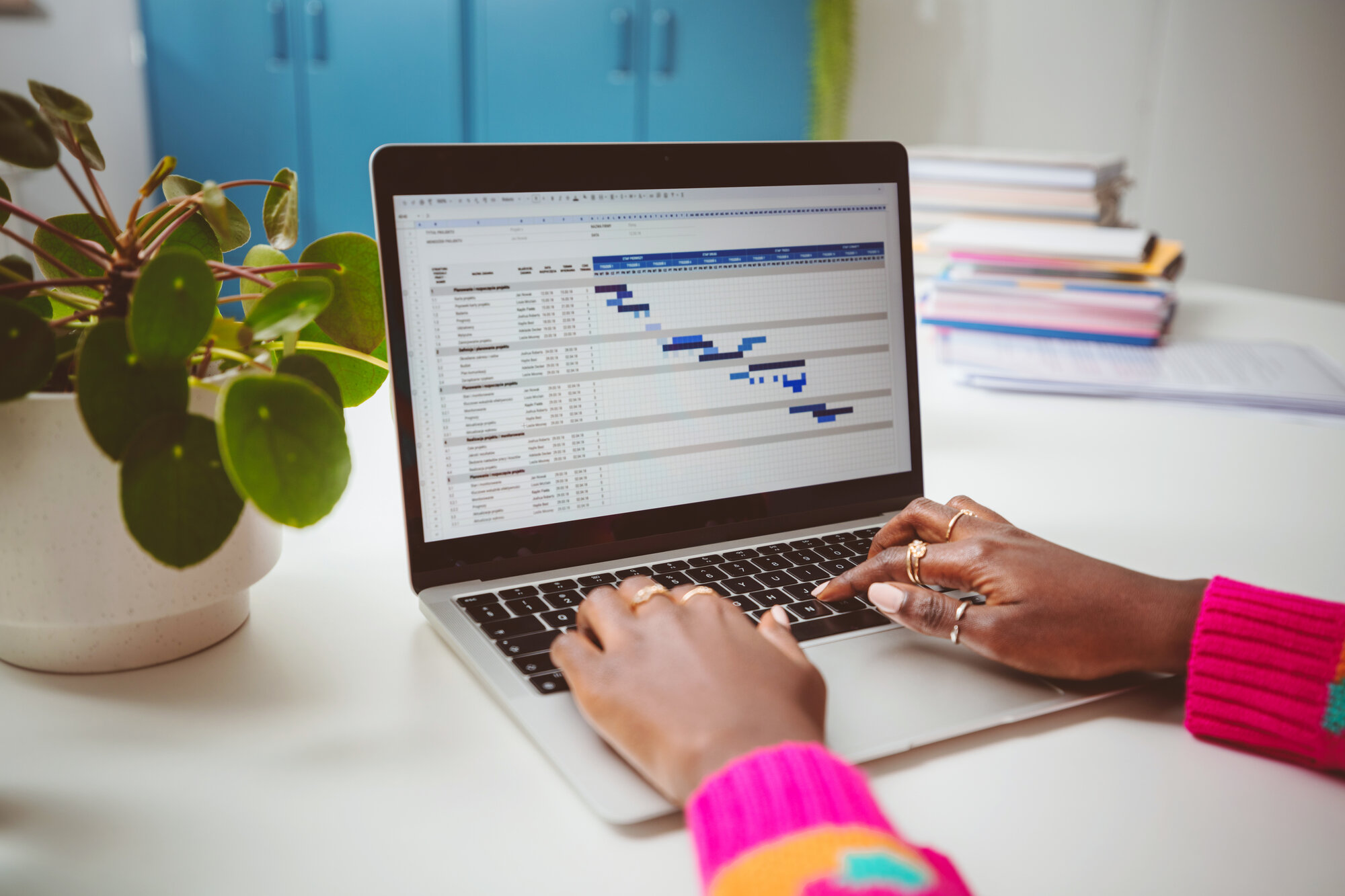 Close-up of hands typing on a laptop showing a hiring pipeline spreadsheet, next to a potted plant.