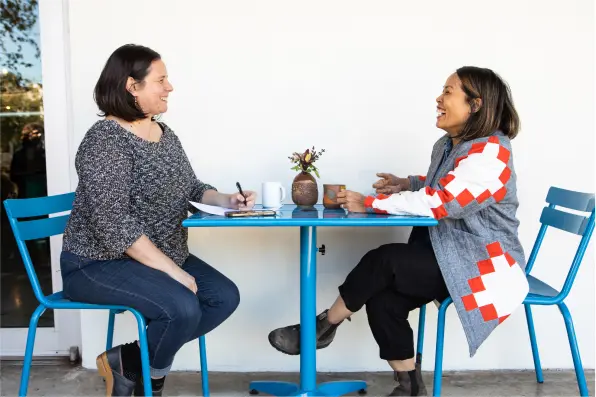 The owner laughs as she sits at a blue cafe table across from another woman who is taking notes with a pen and paper