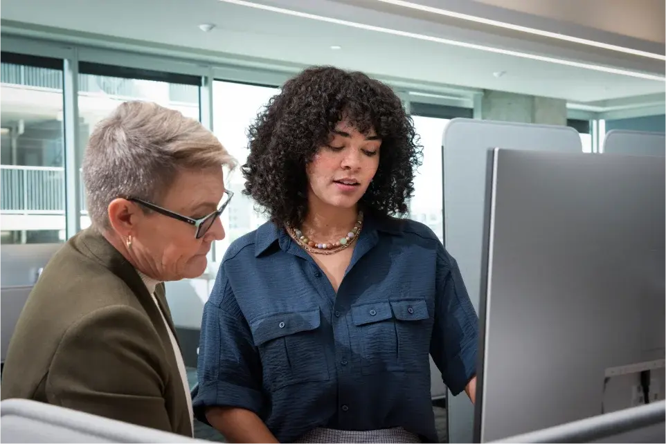 two professionally dressed women of varying ages stand at a desktop computer and discuss candidates for the warehouse positions