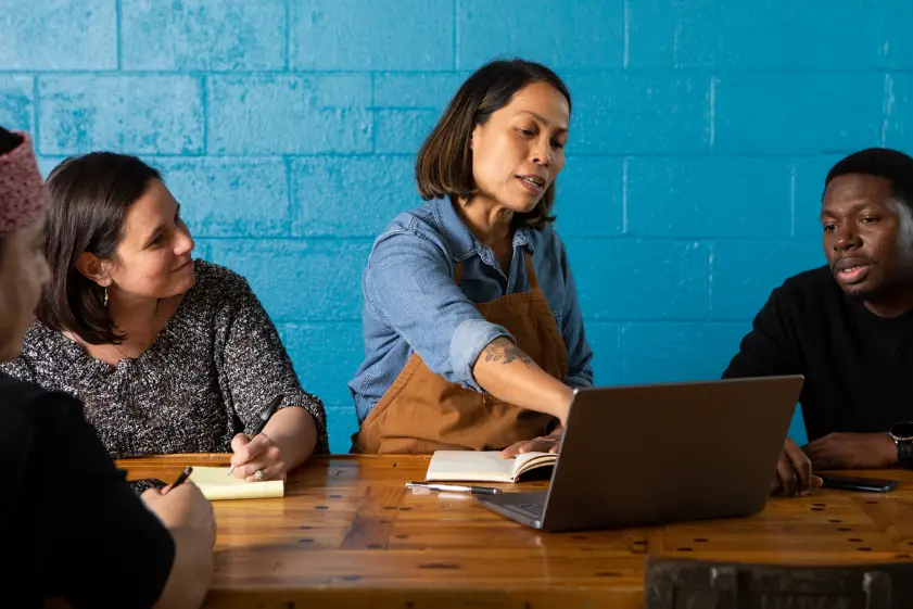 The owner points at her laptop screen and instructs several of her employees who are seated around her