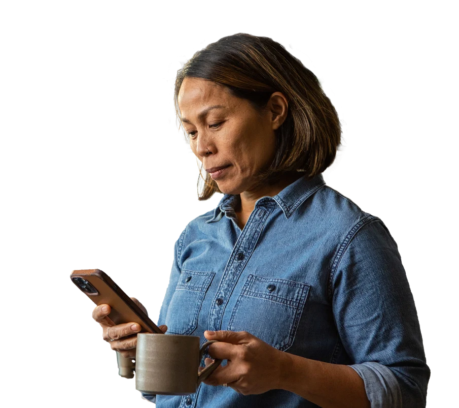 a woman wearing a nose ring and a denim shirt holds a mug as she looks at her mobile phone