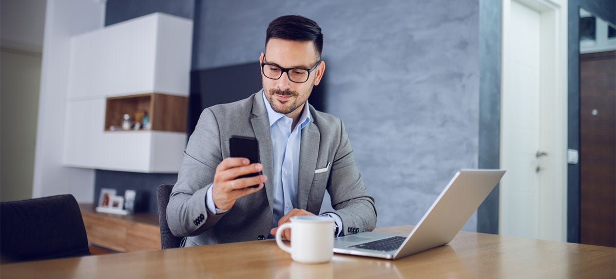 Man checking irregular verbs in English on his phone while sitting in front of a laptop in his office