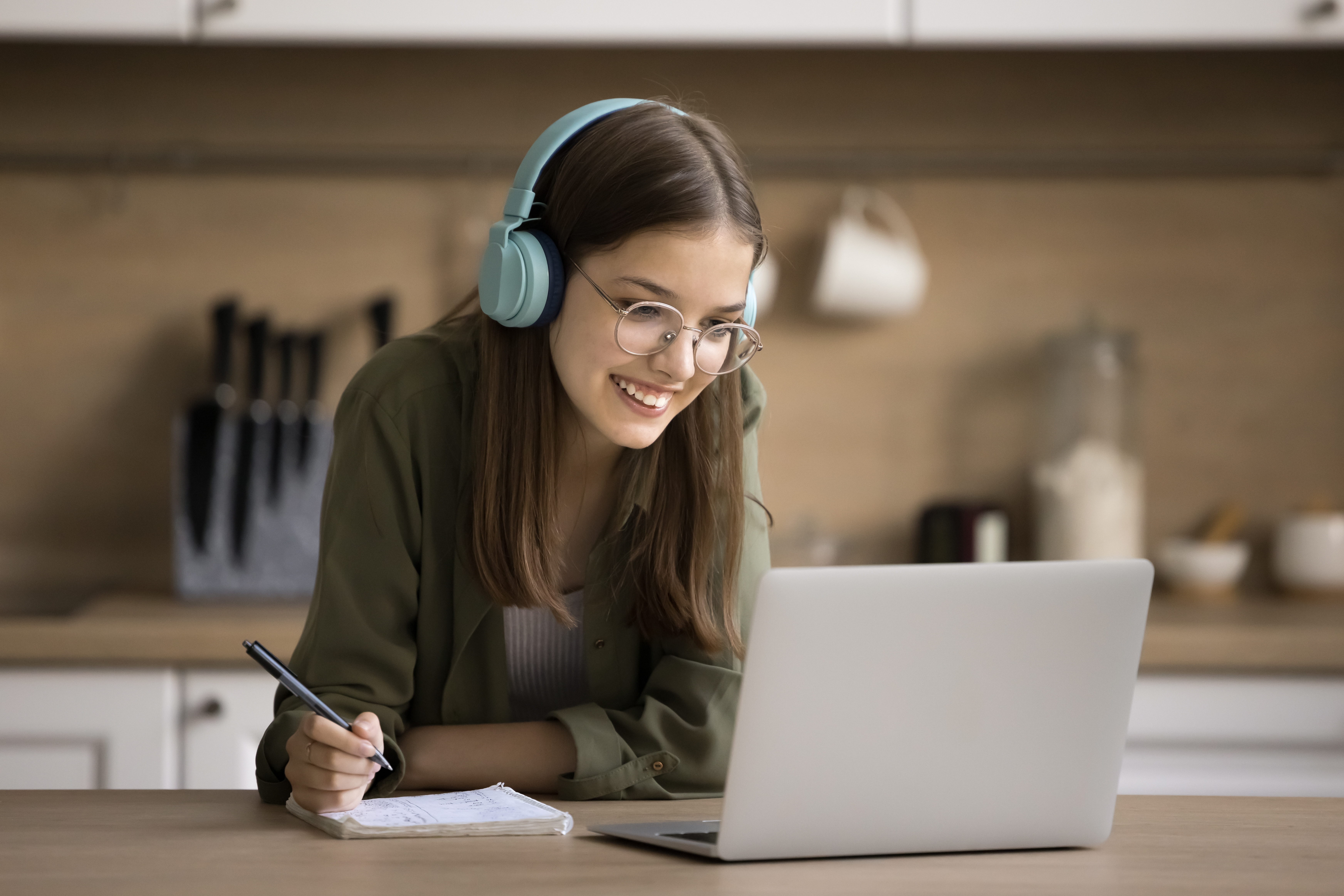 Une fille avec un casque sourit et prend des notes pendant une leçon de langue en ligne pour les enfants et les adolescents avec Berlitz France.
