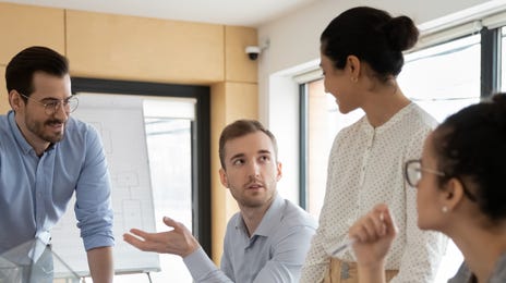 Des collègues de travail se tiennent debout et assis autour d'une table pendant une réunion d'affaires et parlent l'anglais qu'ils ont appris dans un formation anglais entreprise.