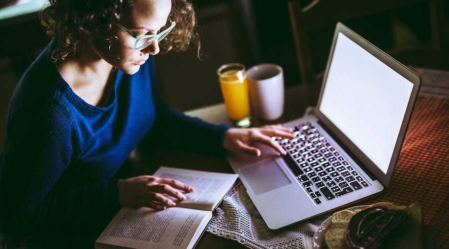 Femme avec un café, assise devant un ordinateur portable et lisant un livre sur les verbes irréguliers en anglais