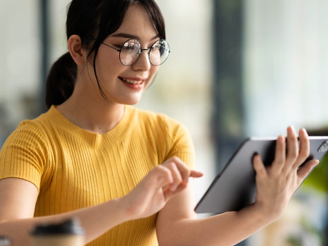 Femme en chemise jaune et lunettes tenant une tablette et s'inscrivant à des cours de langue en ligne avec Berlitz France
