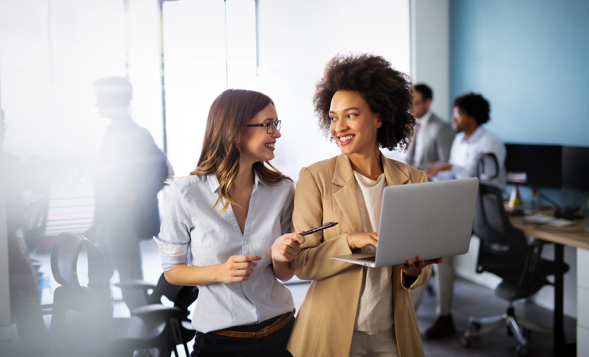 Deux femmes tenant un ordinateur portable et un stylo discutent entre elles après leur cours de langues en ligne pour entreprise.