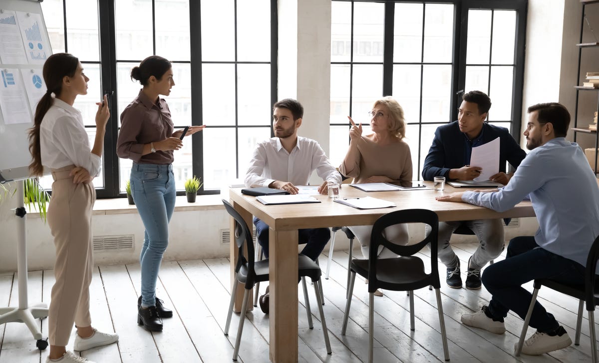 Employés assis autour d'une table et discutant avec leur formateur devant un tableau blanc lors d'un cours de langue des affaires en personne.