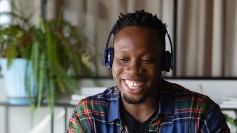 Homme avec des écouteurs prenant des notes et souriant à son professeur lors d'un cours de langue en ligne individuel.