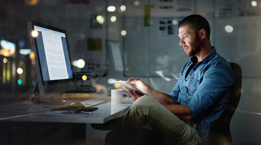 Man in front of a computer holding up a notebook and writing a letter in English there