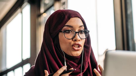 Woman in headphones and scarf sitting in front of her laptop and talking to her instructor during an online English lesson with Berlitz