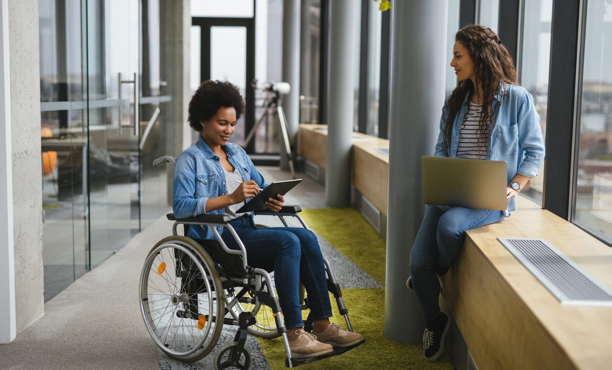 Femme en fauteuil roulant tenant une tablette et discutant avec son professeur de langue dans un couloir avant leur cours d'espagnol pour débutant.