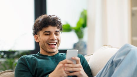 Homme assis sur un canapé, souriant à son téléphone tout en suivant des cours de langue par Skype avec Berlitz France.