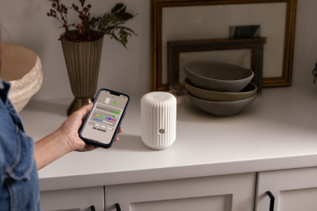 A smart diffuser is featured with a woman's hand holding a smartphone displaying settings from the Scentsy Home app.