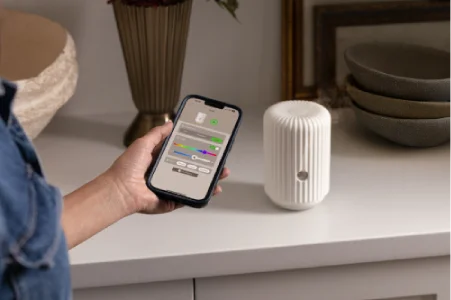 A smart diffuser is featured with a woman's hand holding a smartphone displaying settings from the Scentsy Home app.