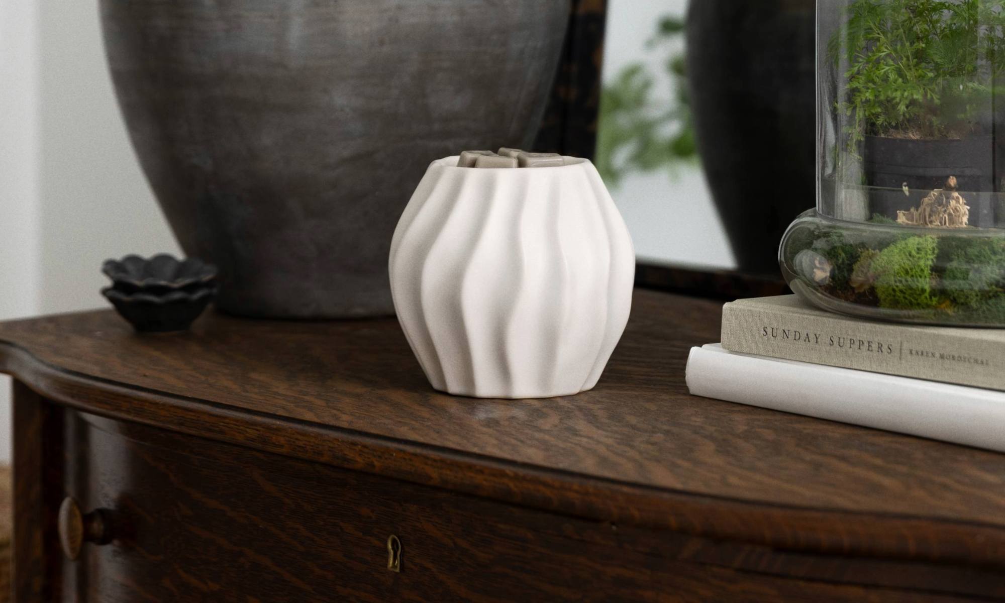 Cascade Warmer in matte white ceramic with soft sculpted curves, styled on a wooden console table beside greenery and decorative accents in a warm, neutral home setting.